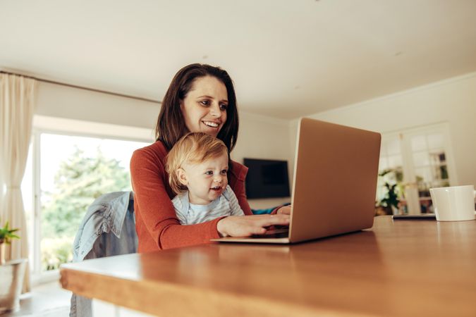 Smiling mother and baby sitting at table and working on laptop