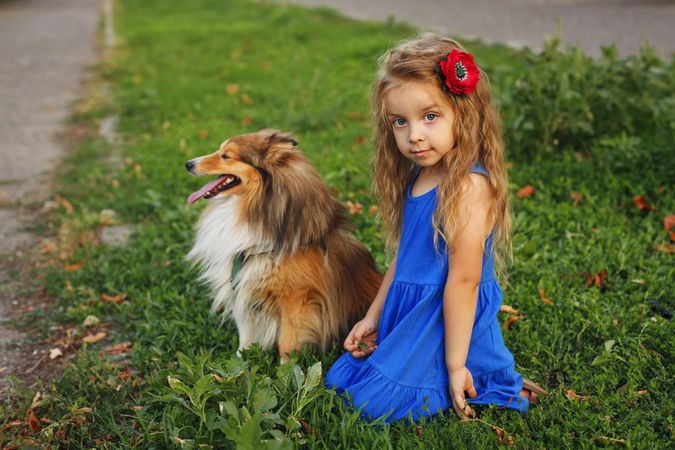 Child in blue dress with flower in hair sitting with dog in the grass