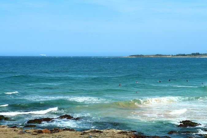 Ocean Waves Crashing Against Rocky Shore With Swimmers In The Blue Sea On A Clear Day