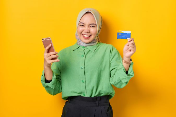 Happy Muslim woman in headscarf and green blouse holding credit card and smart phone up
