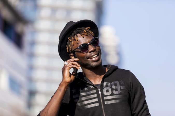 Smiling Black man wearing hat & sunglasses standing on the street talking on cellphone