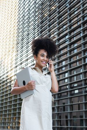 Smiling Black pregnant woman talking on smartphone and carrying a laptop
