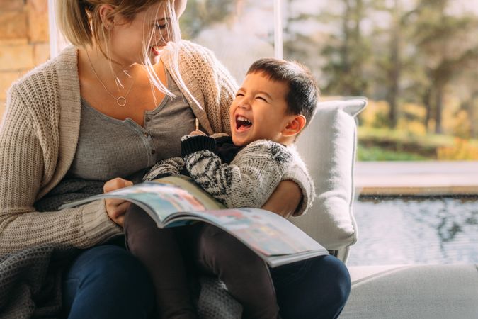 Mother and son looking each other holding a book and laughing while sitting on a couch