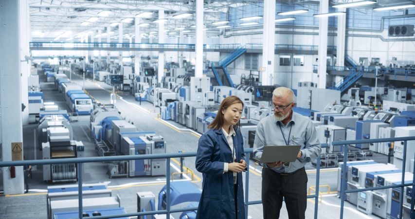 Portrait of a Mature Engineer and Positive Asian Scientist Using Laptop Computer and Talking in a Factory Facility with Equipment Producing Modern Electronic Components for Different Industries