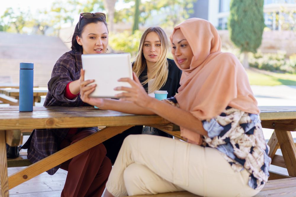 Three women on park bench with insulated mugs watching digital tablet