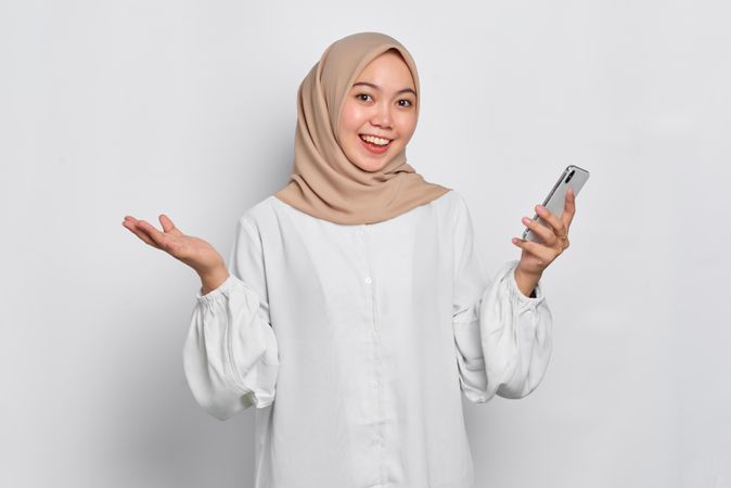Smiling Asian Muslim woman in a bright studio shoot holding up both hands with cell phone in one