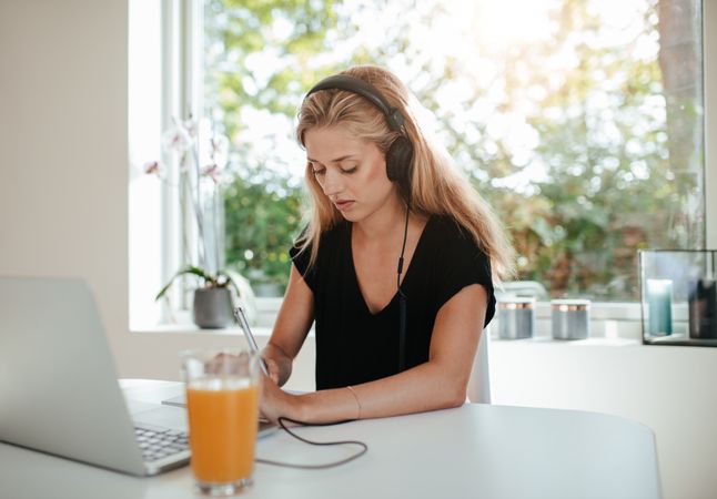 Serious young woman taking notes while sitting in front of laptop computer on kitchen table
