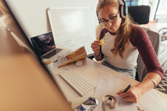Woman writing notes on paper and eating chips at office desk