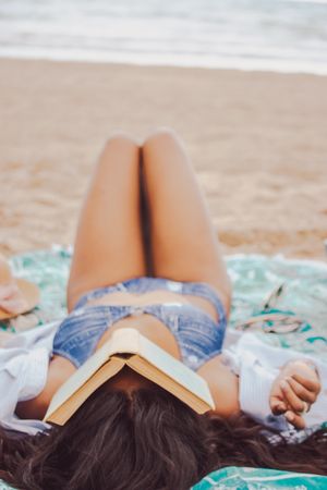 Woman in blue bikini laying on seashore with book on her face