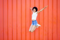 Happy woman in shorts and t-shirt jumping with knees bent against red wall