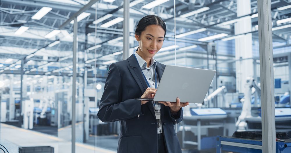 Asian Factory Manager Working on Laptop at Futuristic Industrial Manufacture with Autonomous Robotic Assembly Line. Specialist Monitoring Production, Planning Work for Workers