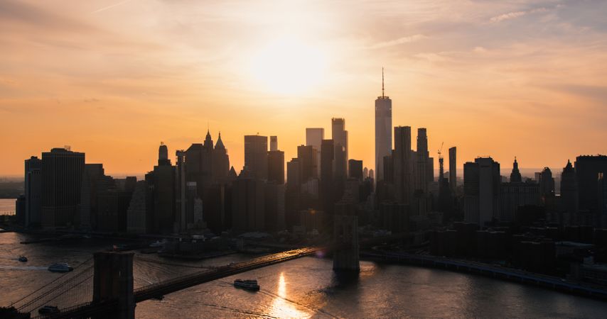Iconic New York City Landscape Over East River with Skyscrapers, Brooklyn Bridge, Cars and Ferry Boats. Cinematic Evening Urban Skyline with Sunset and Lightly Clouded Sky