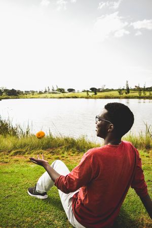 Man in red shirt playing with a ball and sitting on grass field near lake