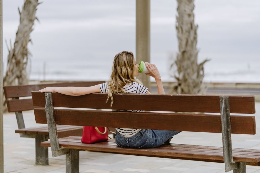 From behind woman with disposable cup of coffee sitting on street bench