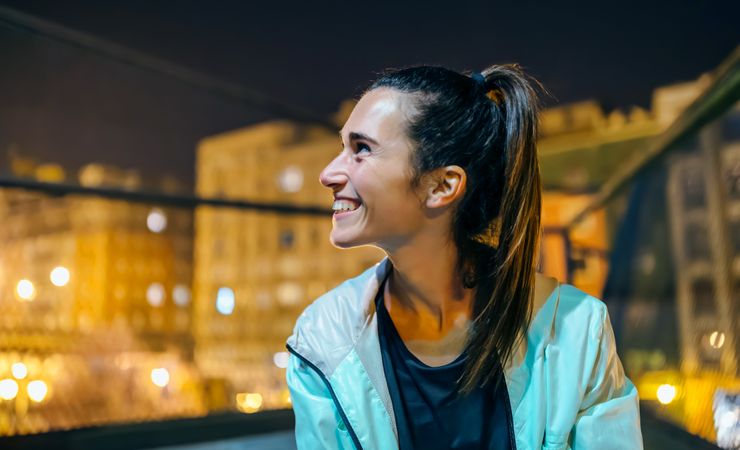 Portrait of cheerful brunette woman with ponytail looking away in city setting at night