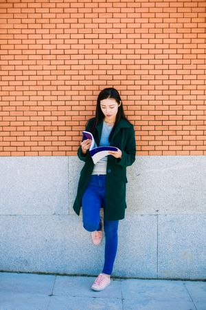 Asian student reading textbook near brick wall