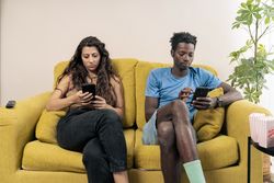 A young couple sits far apart on a couch, each staring silently at their own mobile phone, illustrating digital isolation.