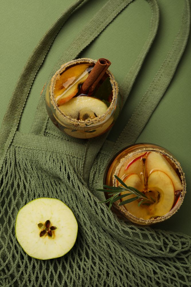 Glasses with apple cider and apple slice on bag on green background, top view