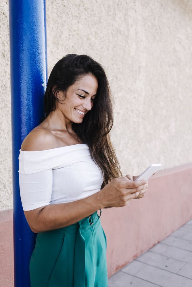 Happy young woman with long hair leaning against wall looking at cell phone