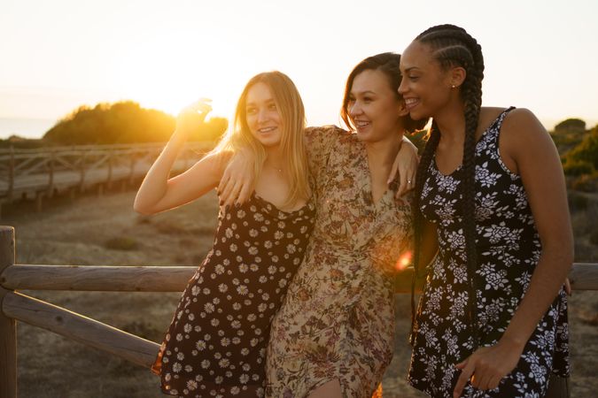 Happy women in summer dresses hanging out together leaning on wooden fence on coast at magic hour
