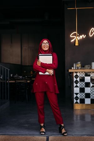 Full length of woman in hijab looking at camera while standing at coffee shop door