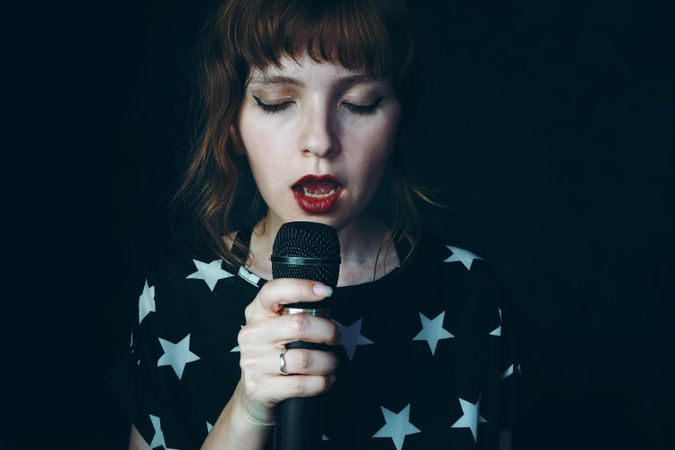 Woman in star print shirt holding microphone singing against dark background