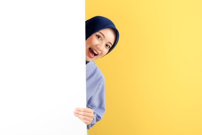 Happy Smiling Young Asian Muslim Woman with Hijab Peeking Out Behind a Wall