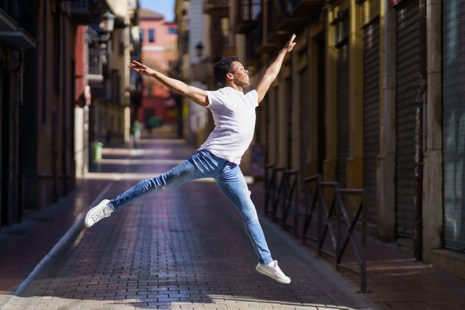 Black male dancer in jeans and t-shirt leaping on an empty European street