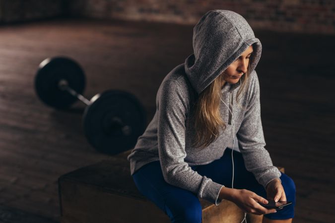 Female wearing a hoodie sitting on a box at gym and looking her mobile phone