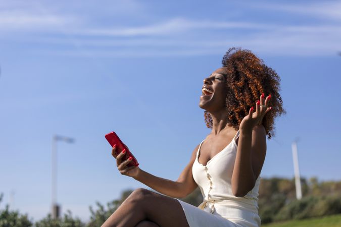 Excited female with curly hair sitting on park grass talking on phone