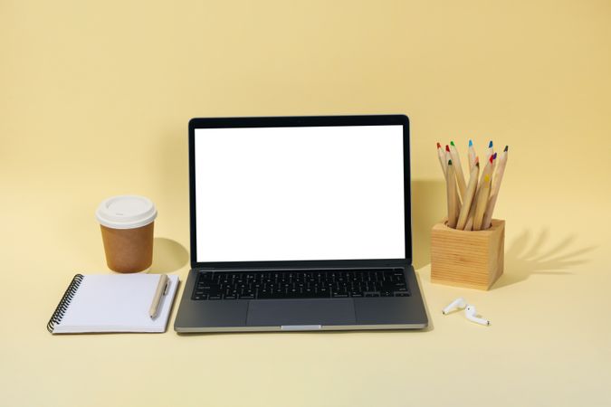 Open laptop with blank mock up screen in cream room with pencil, to go coffee, and notebook with pen