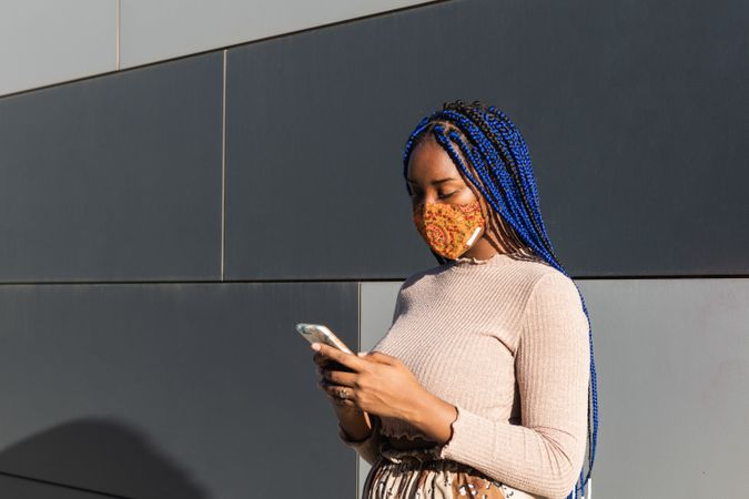 Black woman in mask messaging on smartphone in city