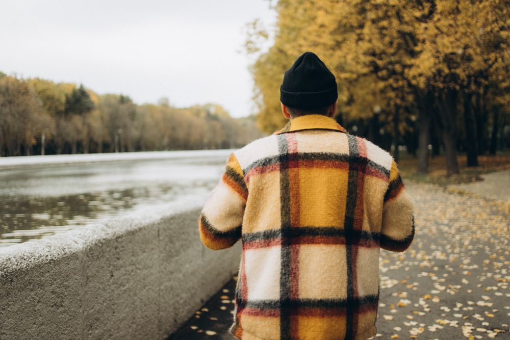 Back view of middle aged man in yellow checkered jacket standing near river and trees