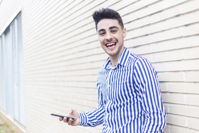 Laughing man leaning on house wall while using his smartphone and looking into the distance