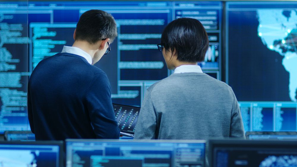 In the System Control Room IT Specialist and Project Engineer Have Discussion, they're surrounded by Multiple Monitors with Graphics.