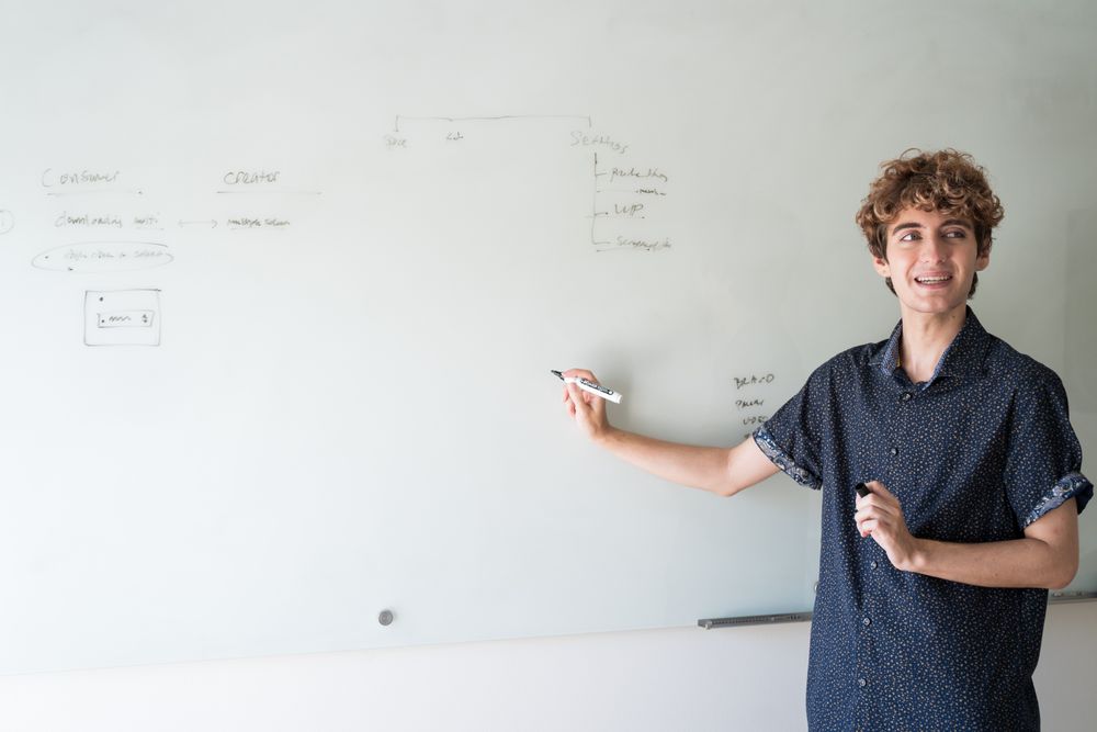 Nonbinary person addressing the room while writing on a dry erase board