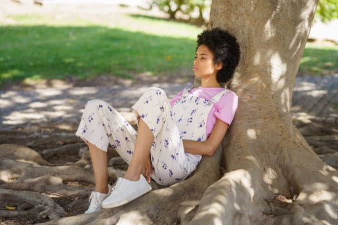 Wistful woman in floral coveralls sitting on roots of large tree in park