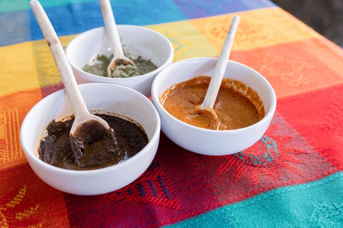 Three bowls with red, green and yellow mole sauce with a wooden spoon on top of a colorful traditional tablecloth