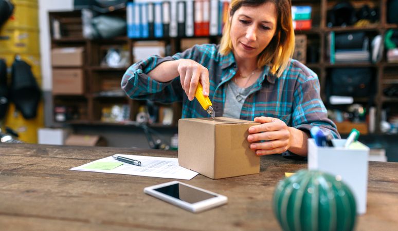 Woman opening cardboard box with cutter in her local store. Parcel delivery and ecommerce concept.