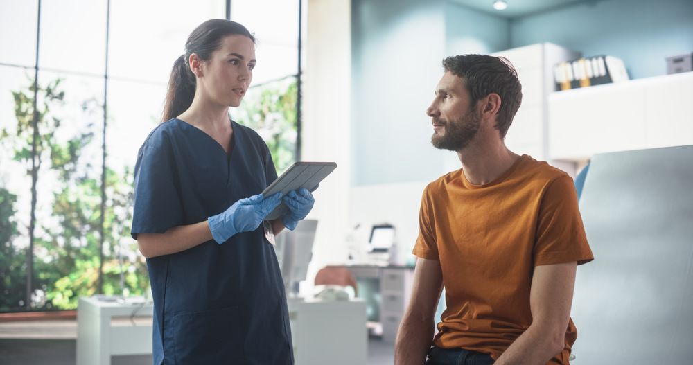 Man Sitting In The Chair In Bright Hospital And Listening To Female Nurse With Tablet Computer.
