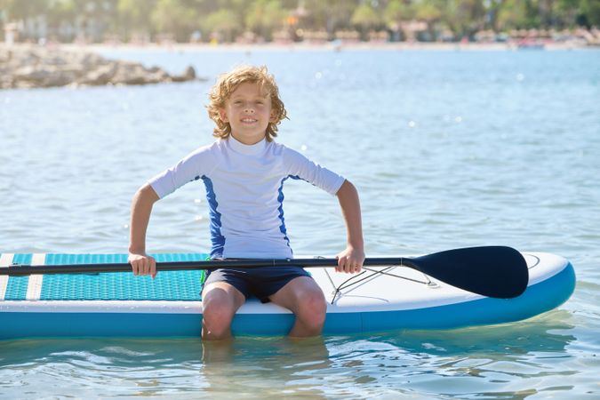 Happy boy in swimwear sitting on paddleboard in water