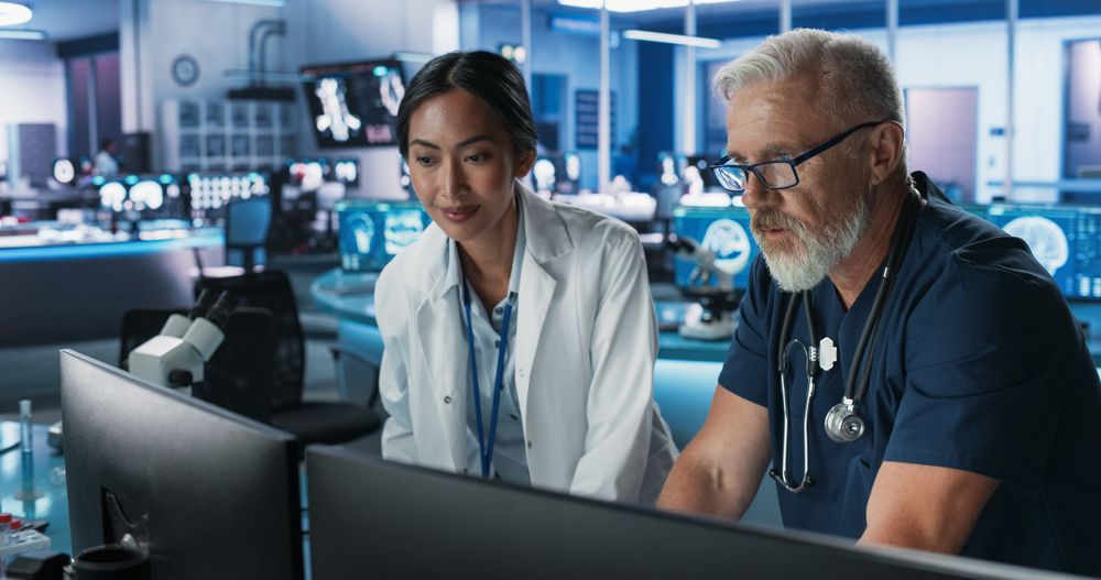 Asian Female Scientist Using Desktop Computer And Talking To White Male Doctor In Medical Hospital Research Lab.