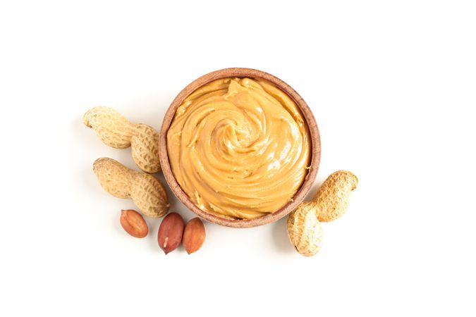 Creamy peanut butter in glass bowl and peanut isolated on light background, top view. A traditional product of American cuisine