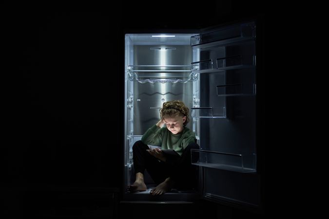 Focused kid watching movie on tablet while sitting in fridge
