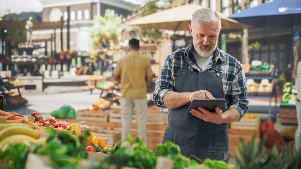 Portrait of a Street Vendor Working on Tablet Computer while Standing in a Farmers Market Stall with Natural Agricultural Products.