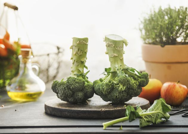 Two whole broccoli heads on kitchen table with ingredients: olive oil, potted herb at window background with natural light. Cooking preparation with healthy green vegetables. Top view.