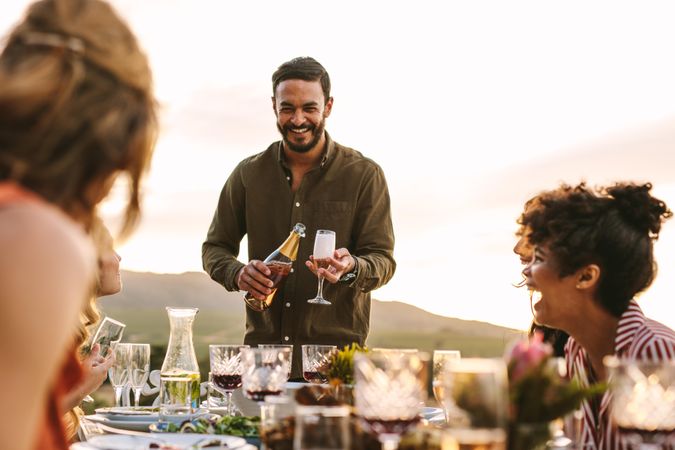 Smiling man serving champagne to friends sitting around table during party