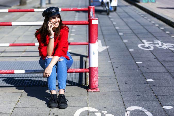 Front view of a beautiful young woman sitting on a metallic fence while using a mobile phone outdoors in the street in a bright day