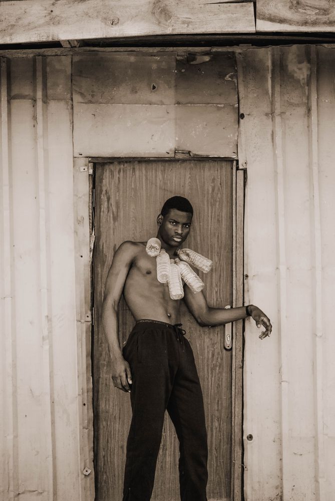 Grayscale portrait of man with a plastic bottles hanging from his neck standing beside door