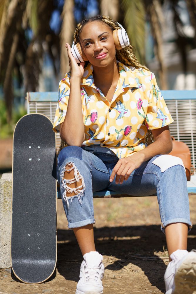 Happy female in bold patterned shirt sitting on park bench listening to music on large headphones